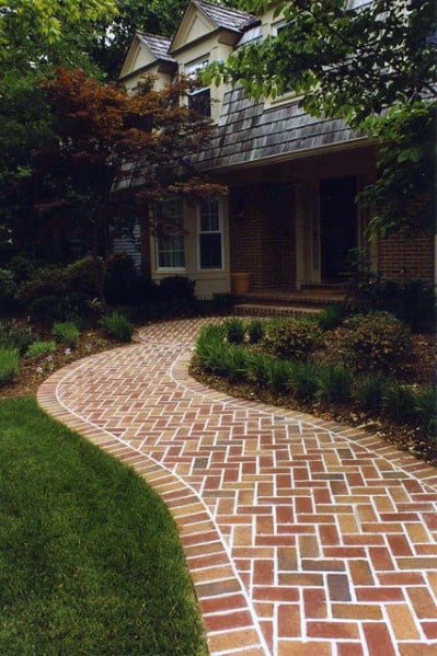Curved brick walkway leads to a brick house with a shingled roof and lush green garden