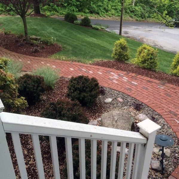 Curved red brick walkway with stone and shrubbery borders, leading through a lush front yard with a view of the street