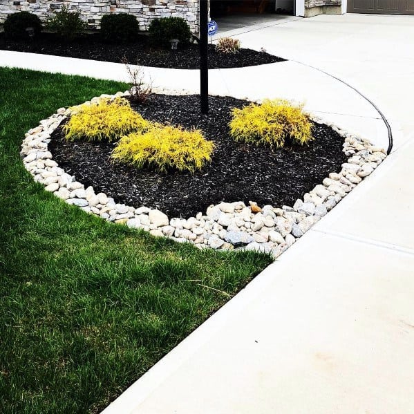A landscaped front yard with a circular stone border around yellow plants and black mulch, featuring a pole light