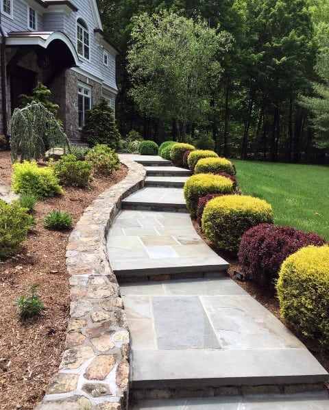 A stone-bordered pathway leading to a house, lined with colorful shrubs and lush greenery on both sides