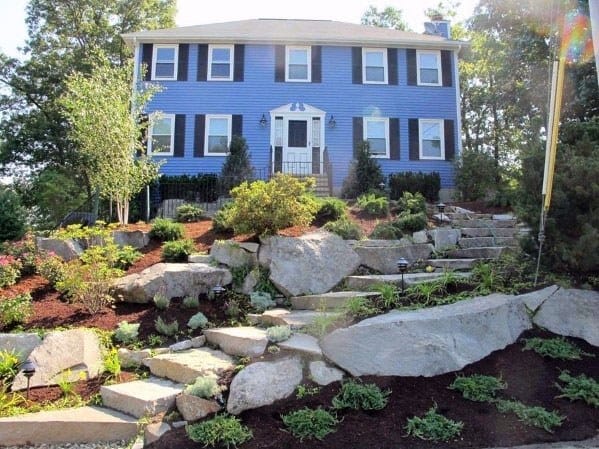 Front yard slope landscaping with stone steps, boulders, shrubs, and greenery leading up to a vibrant blue house
