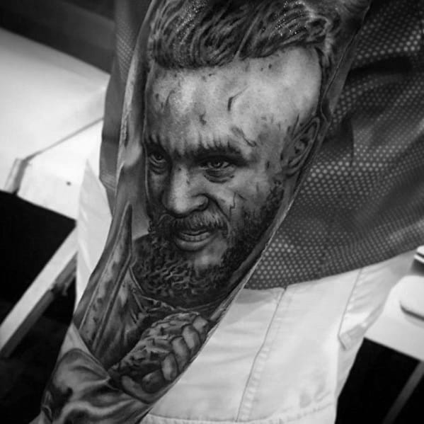 Black and white tattoo of a warrior with a braided hairstyle and beard on an arm