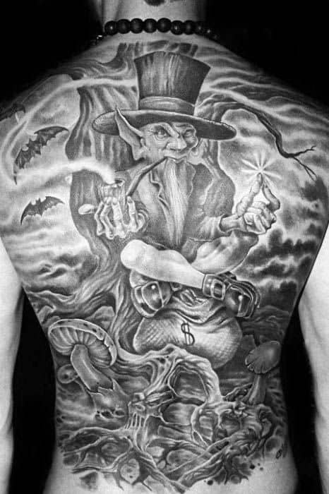 Full back tattoo in black and grey featuring a sinister leprechaun in a top hat holding dynamite and cash, surrounded by bats, skulls, and eerie trees.