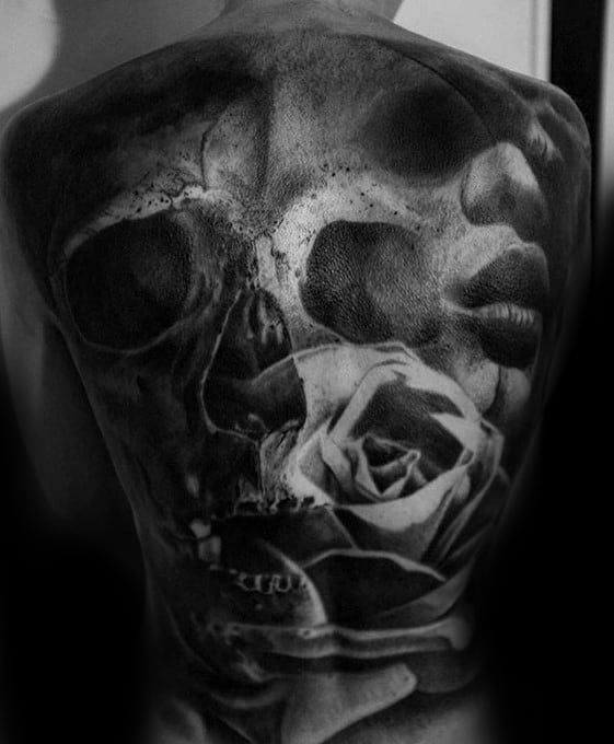 Full back tattoo in black and grey featuring a haunting skull, a delicate rose, and a woman's face blending into the shadows.