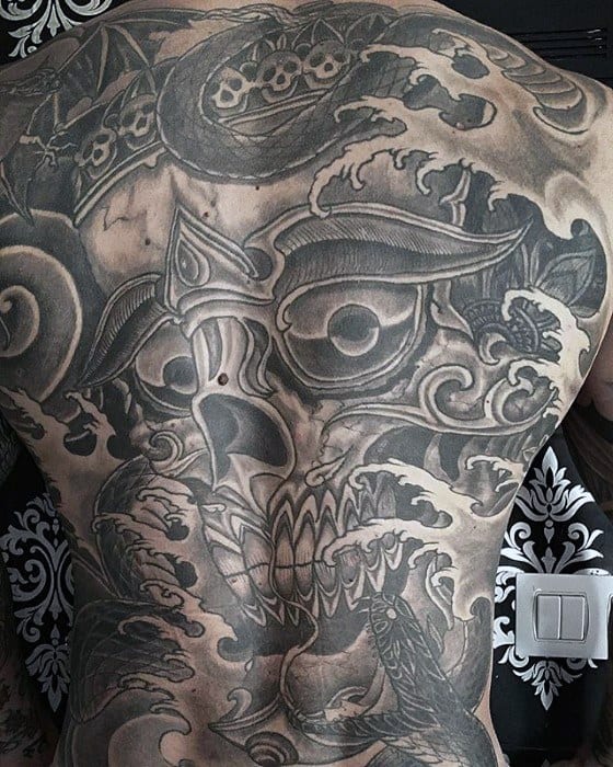 Monochrome Tibetan skull back tattoo with fierce eyes, swirling smoke, and intricate shading covering the entire back.