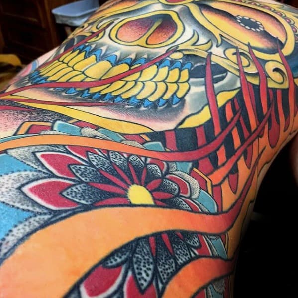 Colorful Tibetan skull tattoo with bold yellow teeth, vibrant floral patterns, and dynamic red and orange wave elements across the back.