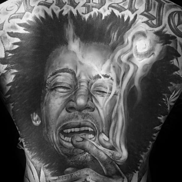 Intense black and gray back tattoo of Bob Marley exhaling smoke, with expressive facial detail and bold lettering across the shoulders.