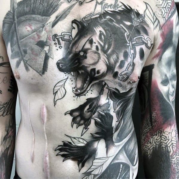 Aggressive black and grey hyena chest tattoo with bold outlines, chains around its neck, and leaves scattered for dynamic movement.