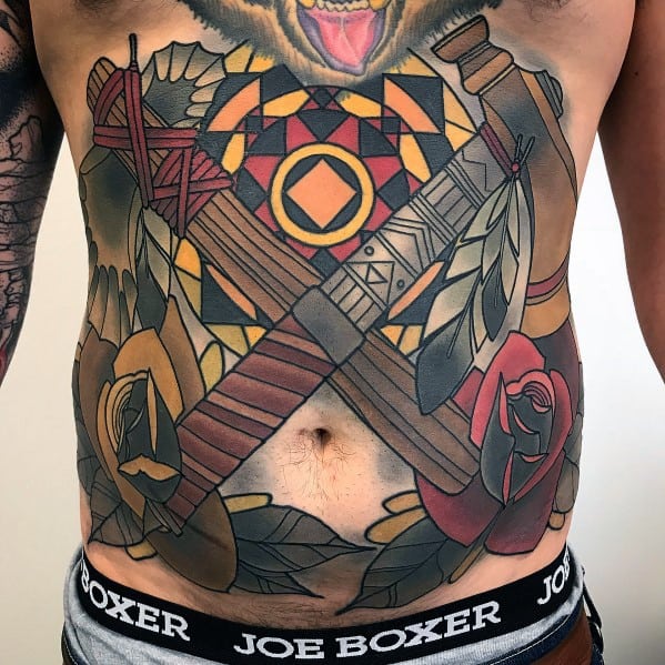 Large chest and stomach tattoo featuring crossed tomahawks with intricate tribal patterns, feathers, roses, and geometric designs in bold colors.