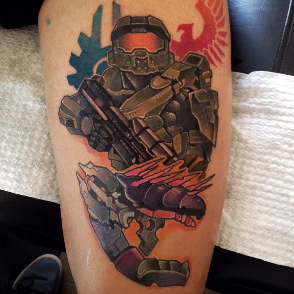 Bold and colorful Halo tattoo of Master Chief wielding a Needler, with UNSC and Covenant emblems in the background, inked on the thigh.