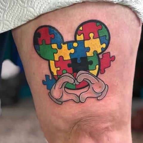 Full color thigh tattoo of Mickey Mouse silhouette with Autism Awareness Puzzle and heart-shaped hands.