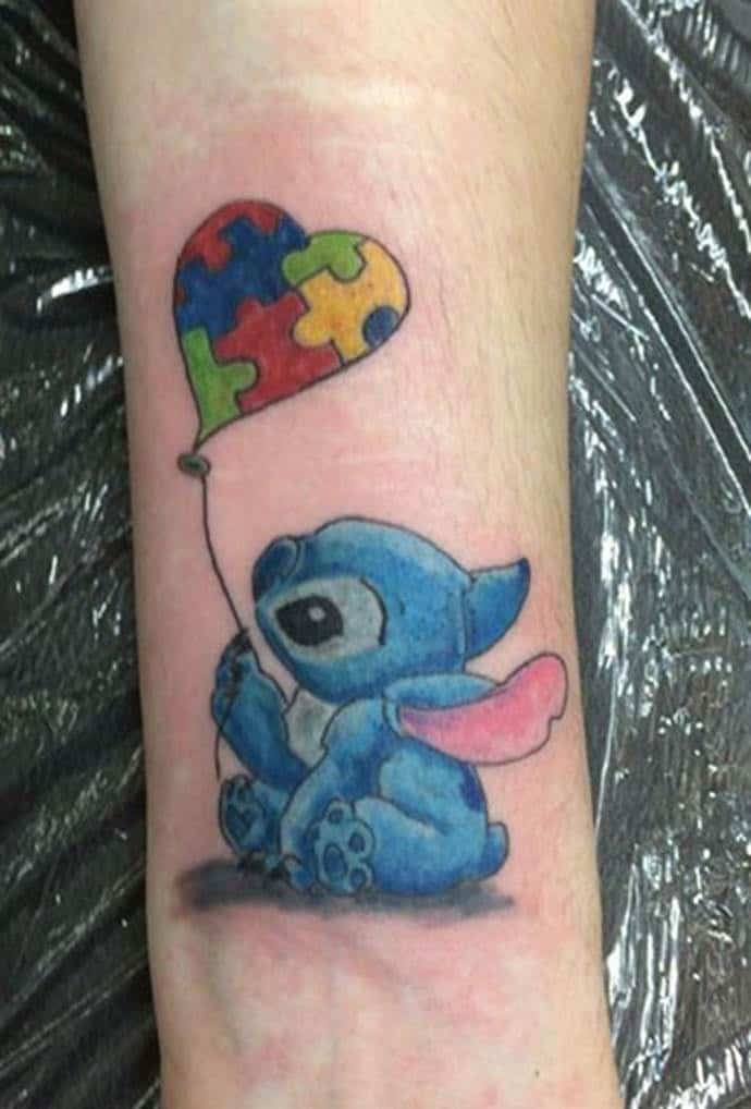 Full color wrist tattoo of Disney character “Stitch” holding a heart shaped, Autism Awareness Puzzle balloon.