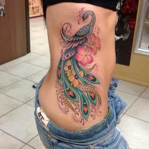 Large and colorful peacock tattoo on the side of the torso, featuring intricate feather details, floral accents, and vibrant blue, green, and gold hues.