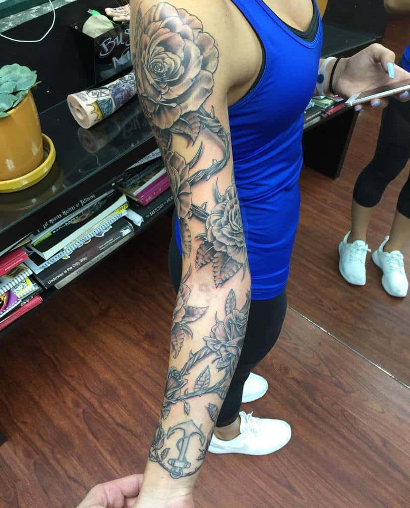 A full sleeve tattoo featuring large, detailed roses intertwined with leaves, accompanied by an anchor design, all in black and gray shading for a striking contrast