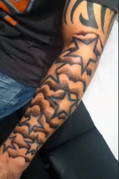 Arm with star tattoos in black ink, surrounded by shaded clouds and abstract designs