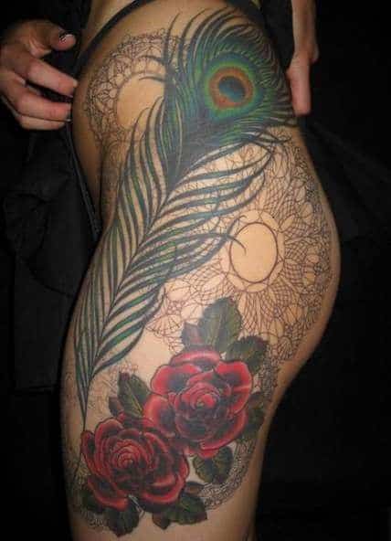 Large thigh and hip tattoo featuring a detailed peacock feather, vibrant red roses, and intricate lace-like mandala patterns for an elegant look.