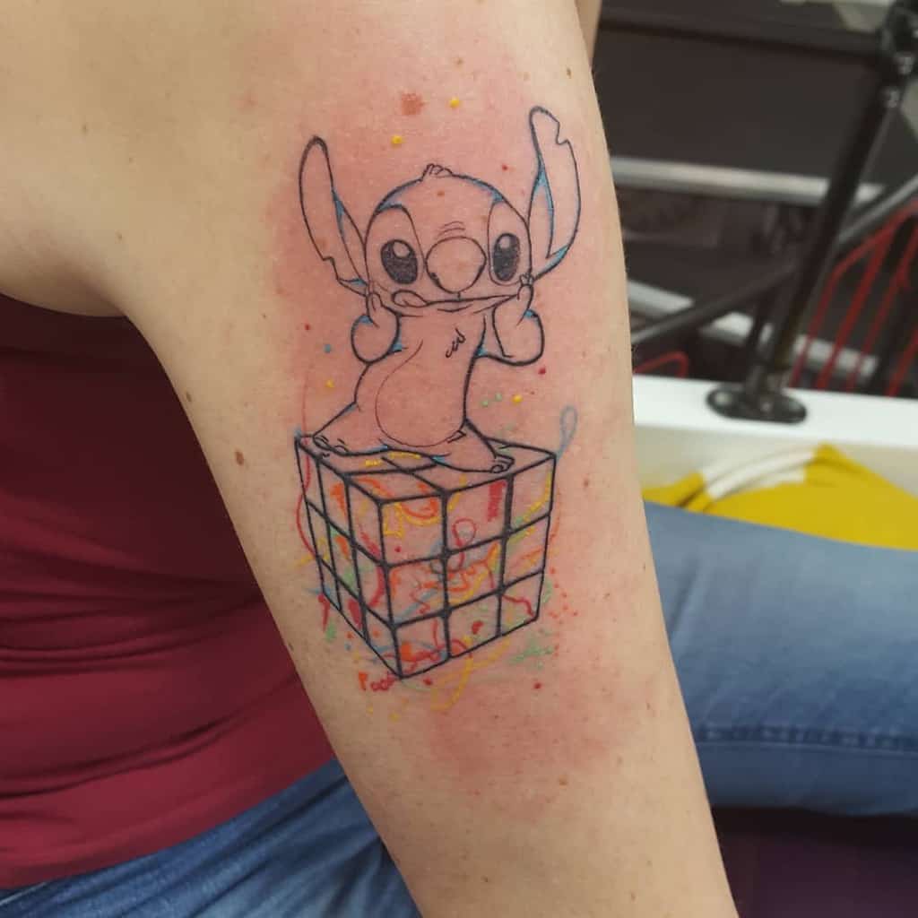 Outline tattoo of Stitch from Lilo & Stitch standing on a Rubik's Cube, with colorful paint splashes, on the upper arm.