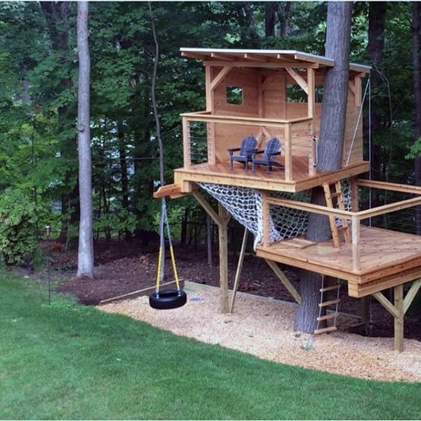 A wooden treehouse with a tire swing, netting, ladder, and chairs, set in a lush forest