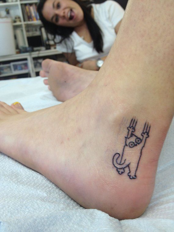A small black ink tattoo of a cartoon cat clinging onto the skin with claw marks, placed on the ankle for a playful effect.