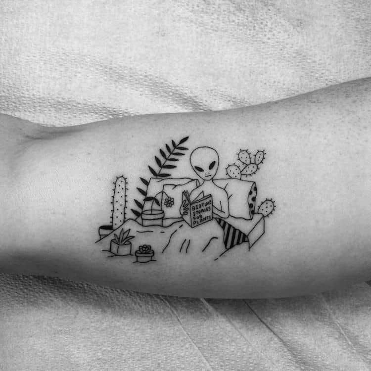 A black ink tattoo of an alien lounging in bed, reading a book titled 'Bottoms Up & the Devil Laughs,' surrounded by cacti and plants.