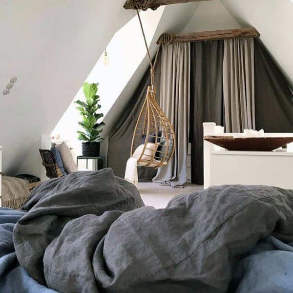 Cozy attic retreat with a hanging chair, lush plants, and soft bedding against slanted walls and large windows