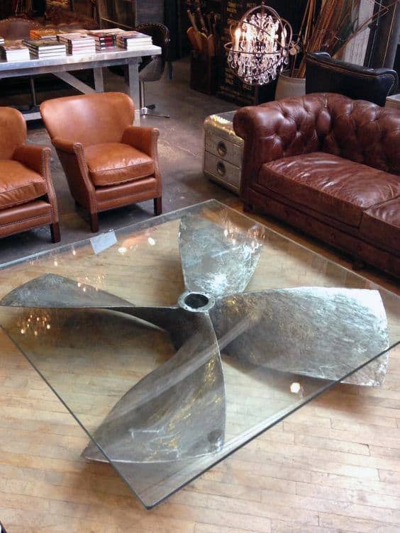 A glass coffee table with a large metal propeller base, surrounded by leather chairs and a sofa