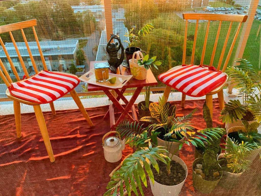Small balcony with wooden chairs, striped cushions, potted plants, and a small table at sunset.
