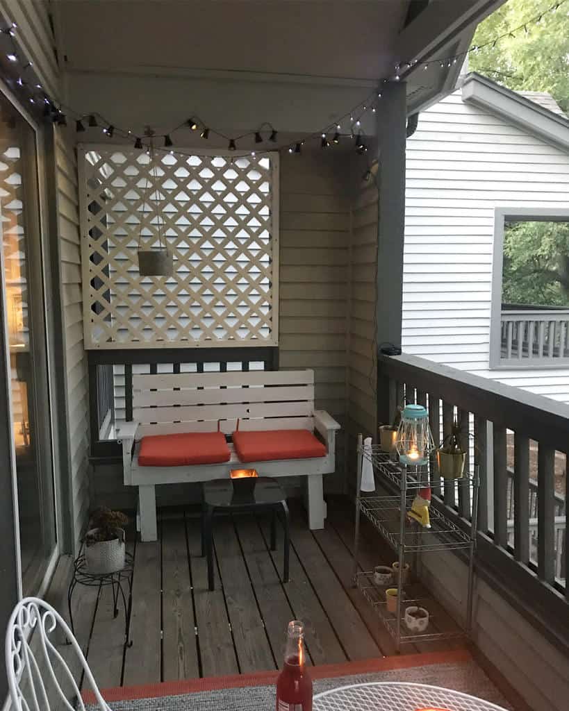 Cozy balcony with white wooden bench, orange cushions, string lights, and small side tables.