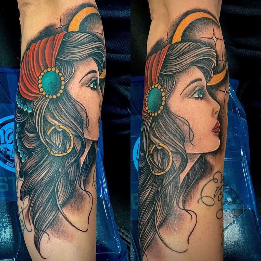 Vibrant tattoo of a gypsy woman with a red headscarf, gold jewelry, and a crescent moon, featuring detailed shading on the forearm.