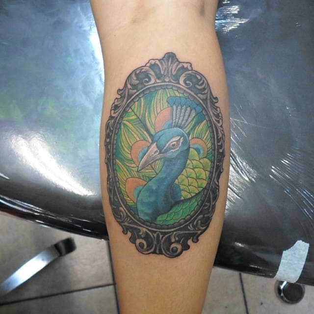 Vibrant peacock tattoo framed in an ornate black border, with colorful feathers in shades of blue, green, and orange on the forearm.