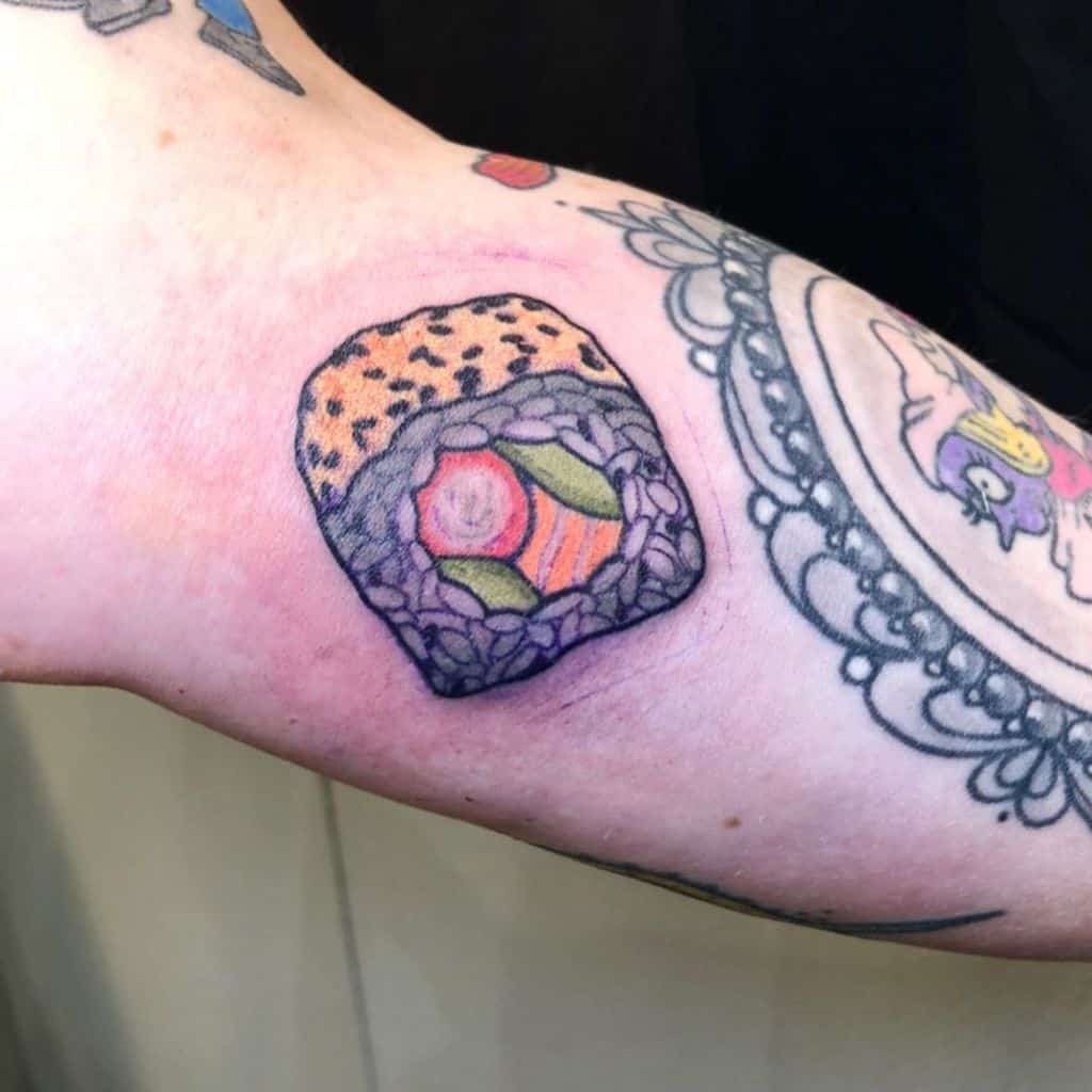 A colorful tattoo of a detailed sushi roll with rice, salmon, avocado, and sesame seeds, placed on the arm in a realistic and playful style.