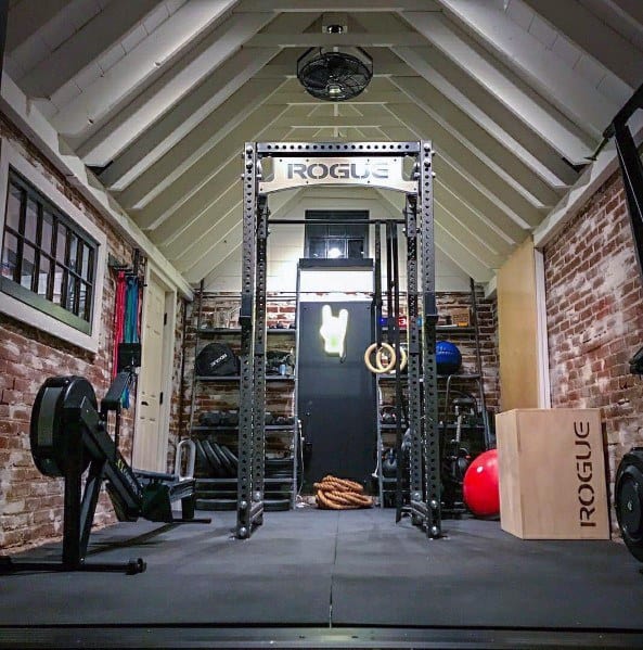 Compact garage gym with a Rogue squat rack, pull-up rings, rowing machine, medicine balls, and plyo box on rubber flooring.