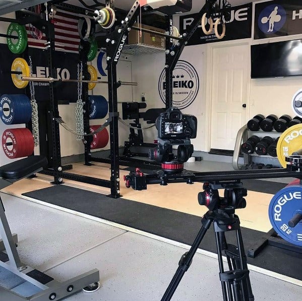 Garage gym with weightlifting equipment, power rack, camera setup, and colorful plates.