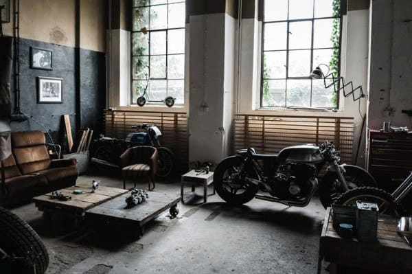 Vintage-style garage workshop with motorcycles, a brown sofa, wooden tables, and large windows letting in natural light