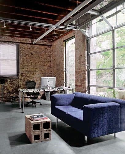 Loft-style office with exposed brick, a blue couch, wooden block table, and garage-like desk by a large window