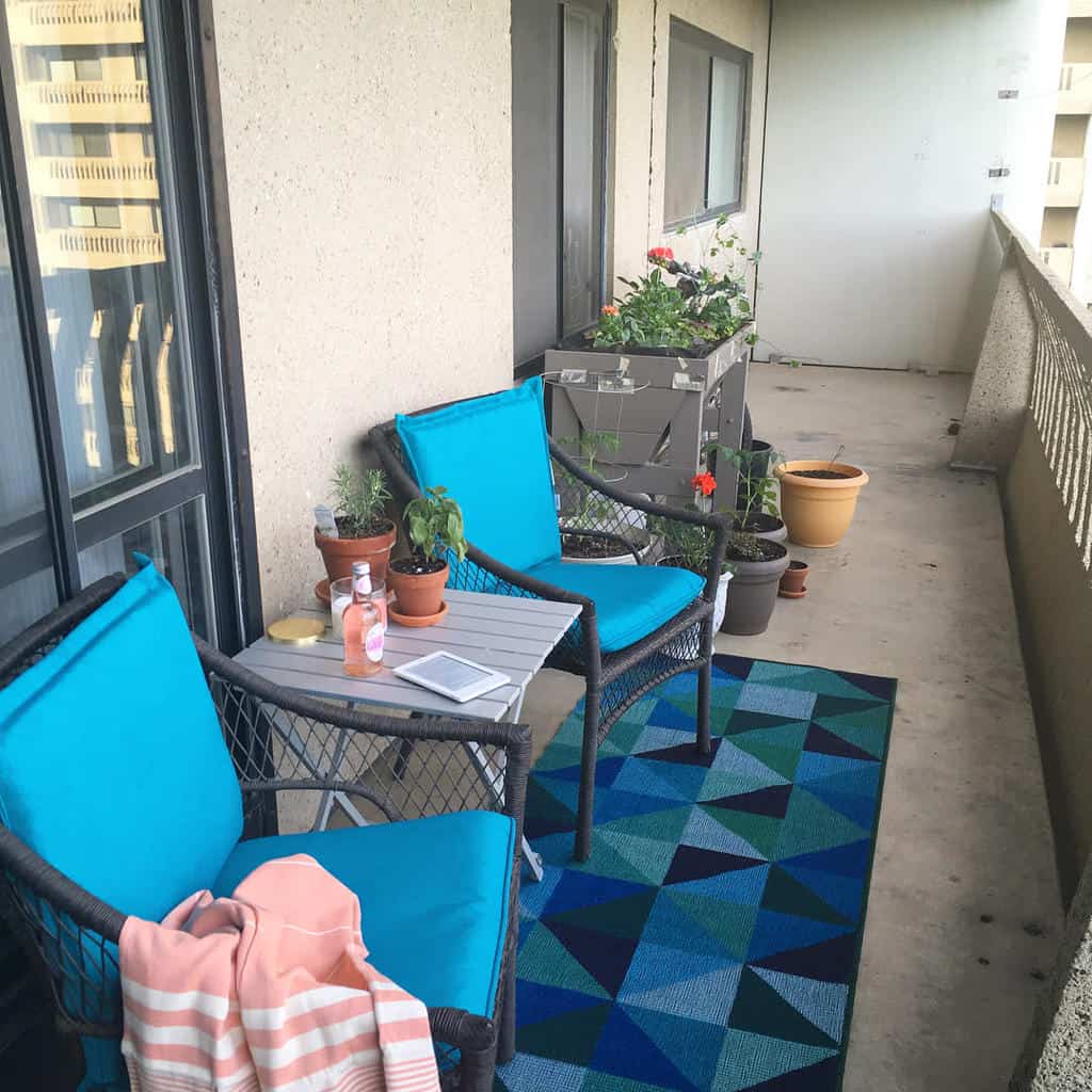 Simple balcony with blue cushioned chairs, potted plants, small table, and geometric rug.