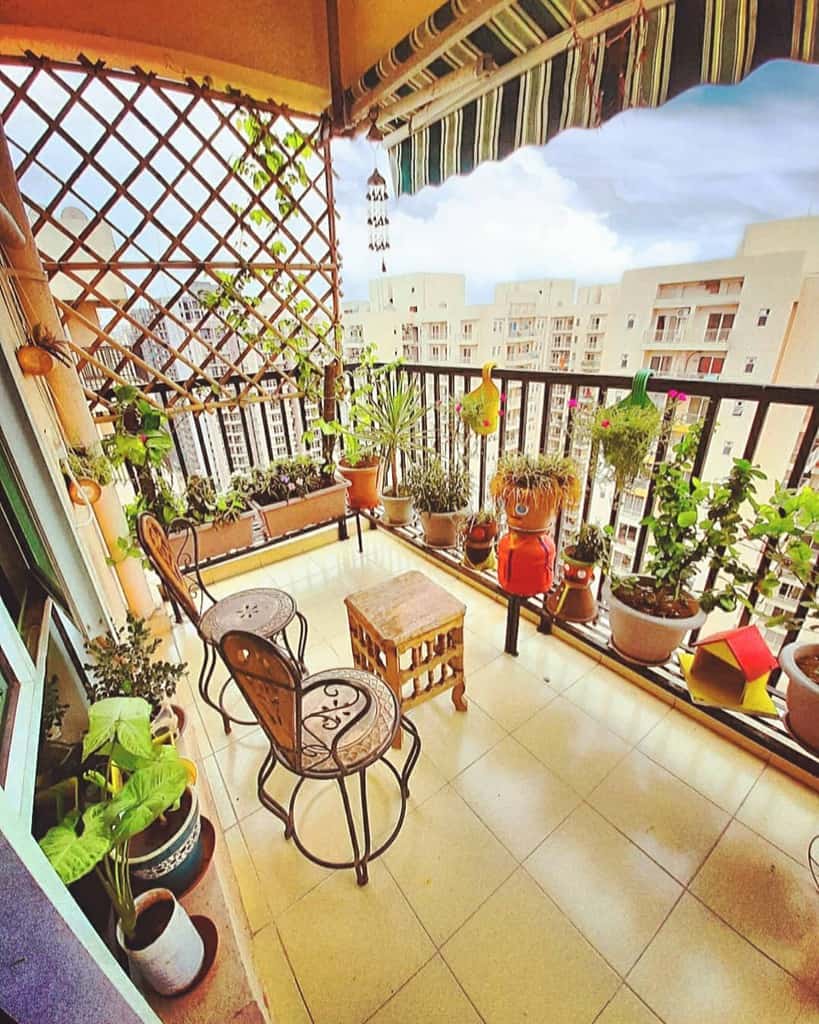 Balcony patio with potted plants, metal chairs, a wooden stool, and a trellis against the railing.