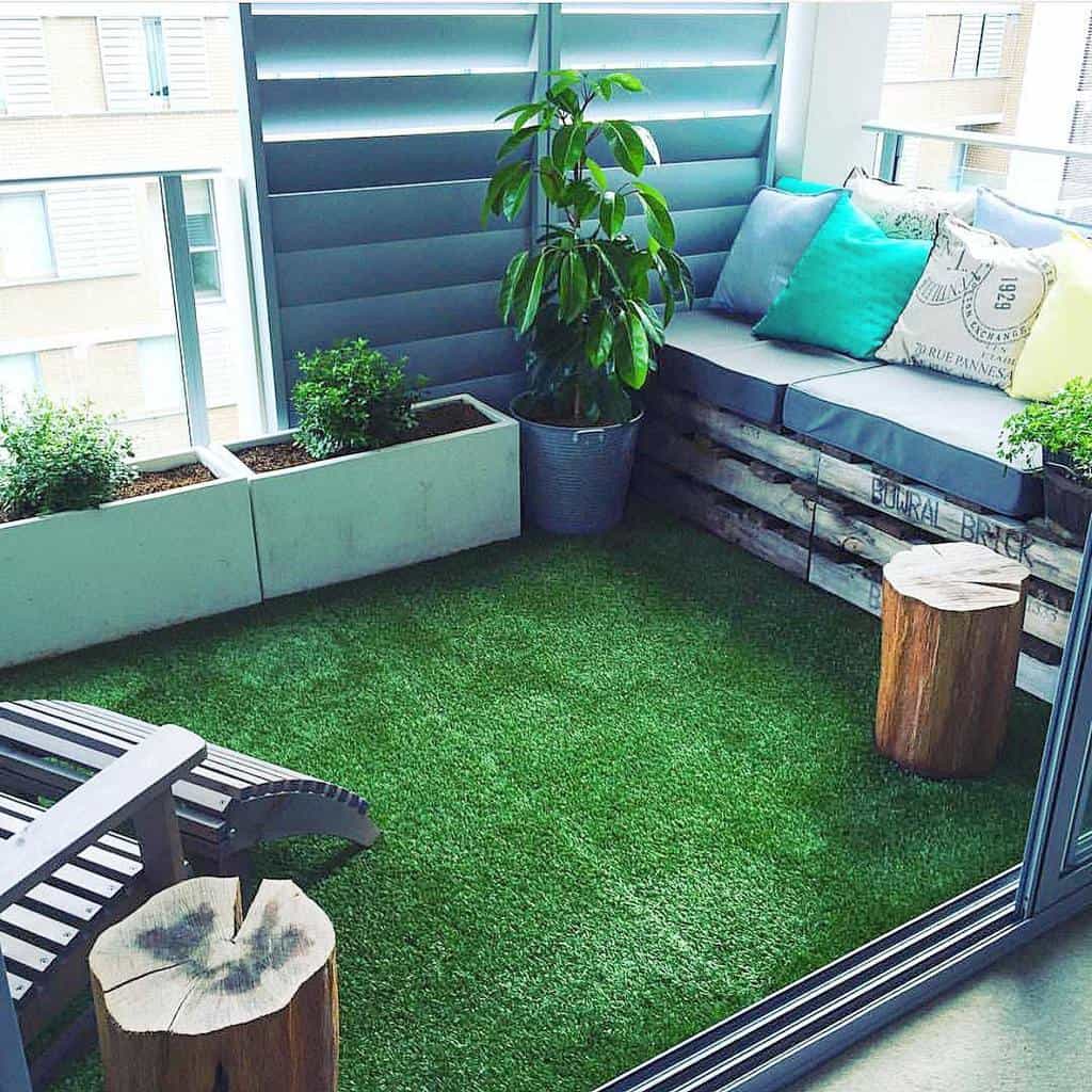 Small balcony with artificial grass, wooden seating, potted plants, and cozy cushions.