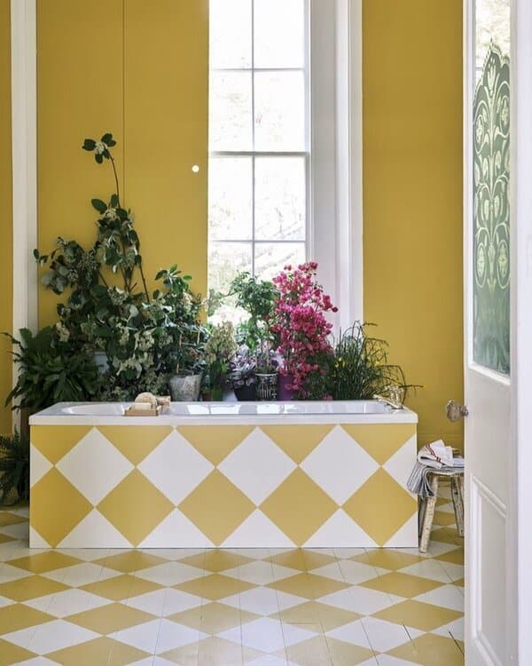 Bright yellow bathroom with a harlequin-patterned tub surround, lush plants by a tall window, and diamond-patterned flooring.