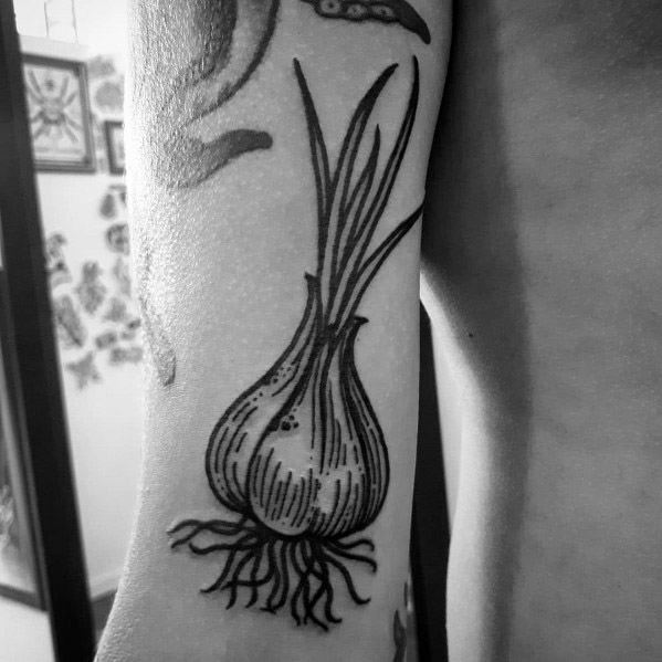 Bold black ink garlic tattoo on the arm with fine line details, long stems, and roots, creating a striking botanical design.