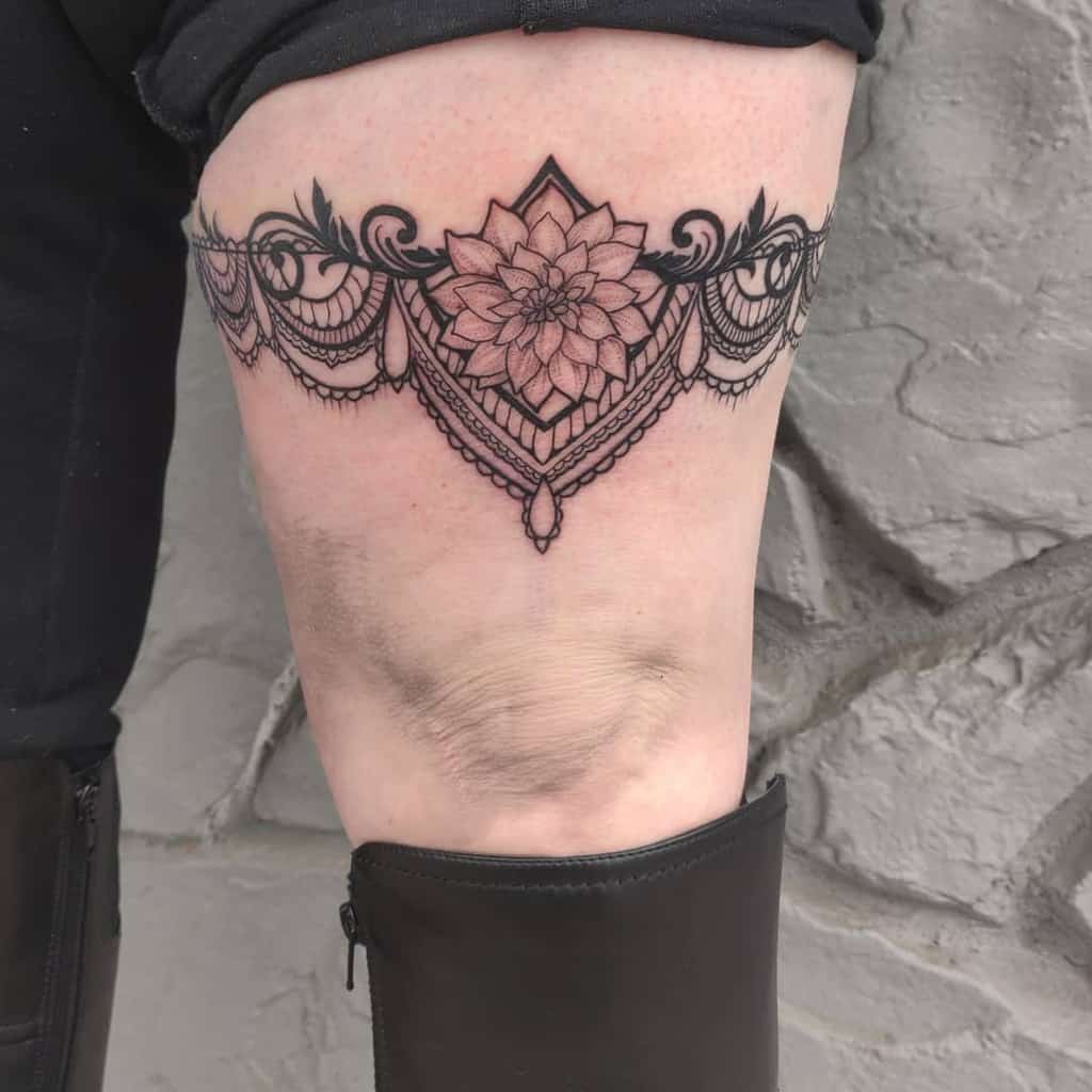 Elegant thigh tattoo resembling a lace garter, featuring a detailed floral mandala centerpiece with ornamental swirls and beaded accents.