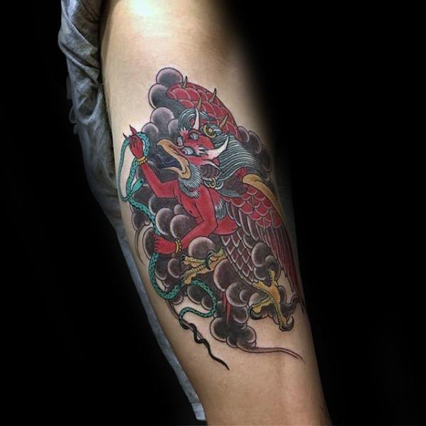 Garuda tattoo on the upper arm featuring a red winged figure holding a green serpent, set against dark clouds with gold and black accents.