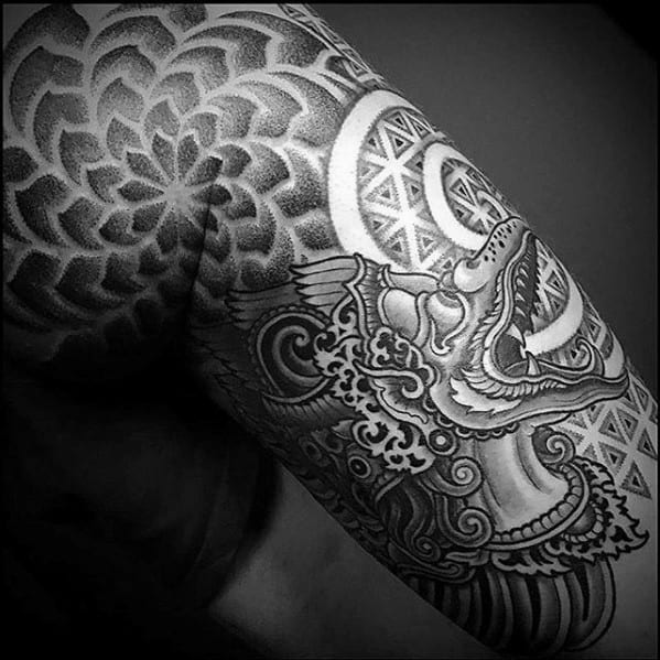 Garuda tattoo on the upper arm with a stylized beaked figure, surrounded by geometric patterns and dotwork creating a bold, sacred design.