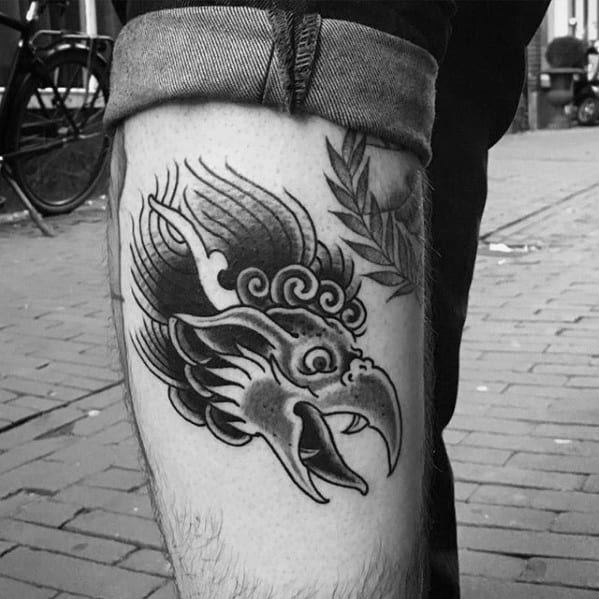 Garuda tattoo on the calf featuring a bold, stylized bird head with sharp beak, flowing lines, and a small leafy branch beside it.