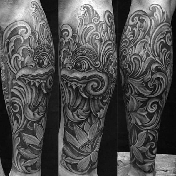 Garuda tattoo on the calf in black and grey, featuring a fierce mythological face surrounded by flowing ornamental patterns and a lotus flower.