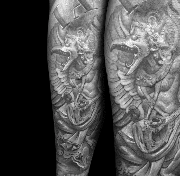 Realistic Garuda tattoo on the forearm in detailed black and grey, featuring a fierce winged figure with sharp claws, expressive face, and layered armor.