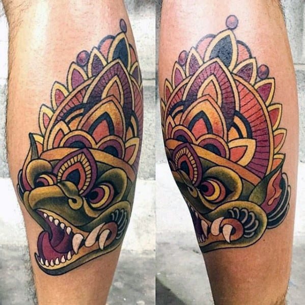 Colorful Garuda tattoo on the calf with a fierce expression and a crown-like headdress in bold shades of red, orange, yellow, and purple.