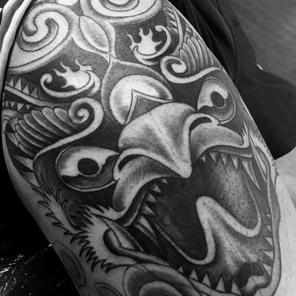 Garuda tattoo design with a bold, open-beaked expression and swirling patterns, covering the upper arm in black and grey ink.