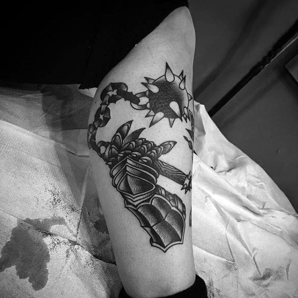 Black and grey tattoo of an armored gauntlet wielding a spiked flail, with bold shading and dynamic chain movement on the upper arm.