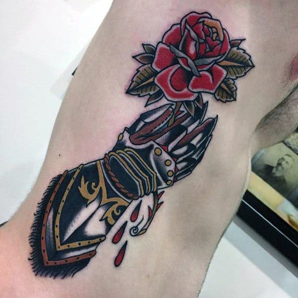 Bold tattoo of an armored gauntlet pierced by a rose, with red petals, blood drops, and gold detailing on the armor.
